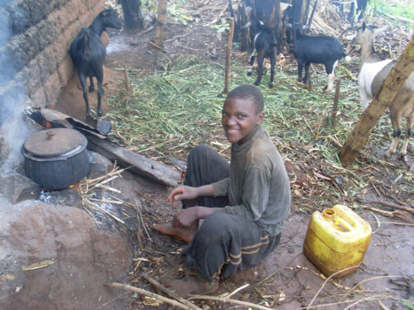 Ruandischer Junge an offenem Herdfeuer mit schwarzen Ziegen im Hintergrund