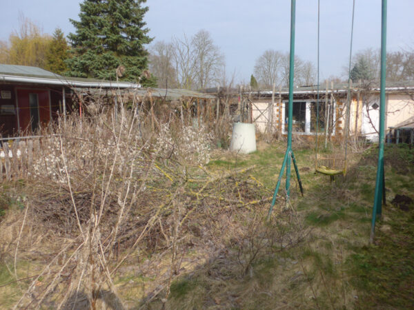 Trockene Pflanzen, abgesägte Äste auf Haufen, Gartenhaus und Tomatenschutzdach vom hinteren Zentralbereich aus gesehen